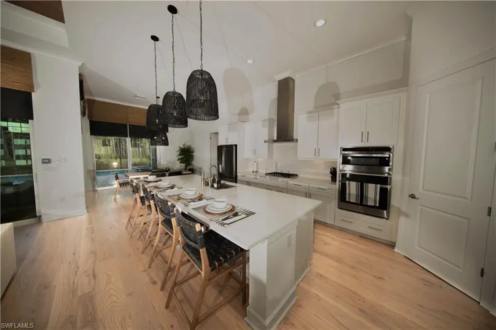 Kitchen with wall chimney exhaust hood, appliances with stainless steel finishes, a large island with sink, light wood-type flooring, and white cabinets