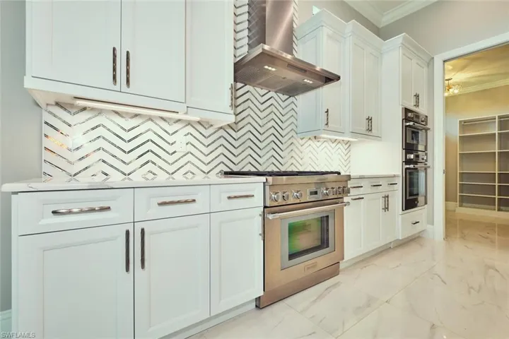Kitchen featuring wall chimney exhaust hood, stainless steel appliances, crown molding, light marble finish flooring, and white cabinets