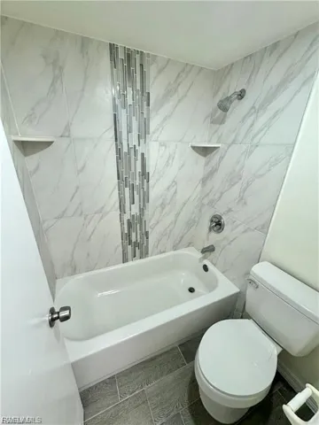 Bathtub with a shower, white subway tile surround, decorative tile inlay, corner shelving, and a toilet