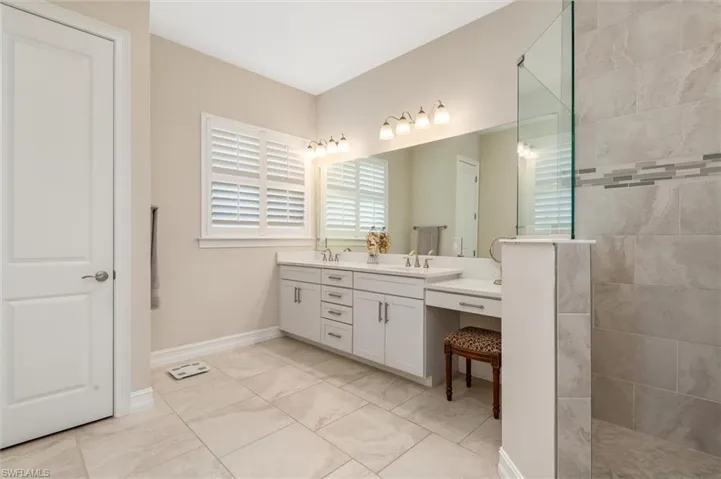 Spacious bathroom featuring large format tile flooring, a dual vanity with light cabinetry and solid surface countertops, a dedicated makeup area, and a walk-in shower with a glass enclosure and decorative tile band