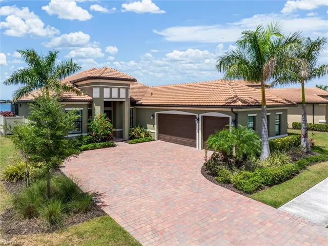 Mediterranean-style residence featuring a terracotta tile roof, a spacious paver driveway, two integrated garages with arched doors, mature palm trees, and lush landscaping