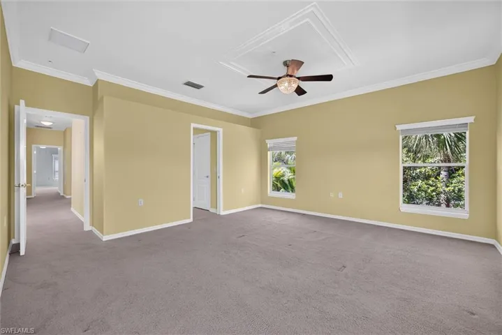 Spacious interior room featuring a tray ceiling with crown molding, a ceiling fan with light fixture, two windows, and neutral carpeting