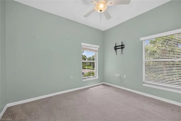Carpeted room featuring two windows with horizontal blinds, a ceiling fan with integrated lighting, and a wall-mounted television bracket