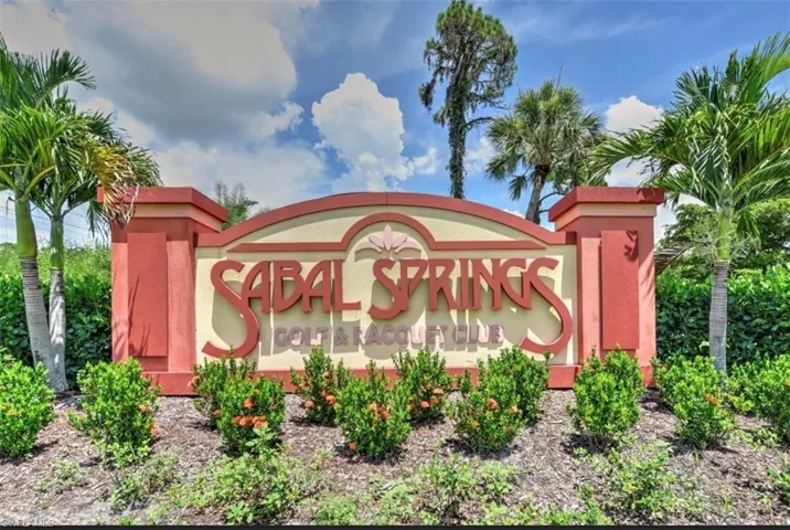 Sabal Springs Golf & Racquet Club entrance sign, featuring multi-toned architectural elements, decorative landscaping with green shrubs, and mature palm trees