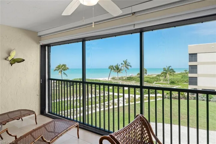 Screened balcony featuring a ceiling fan, tile flooring, and a black metal railing overlooking the beach, ocean, and palm trees