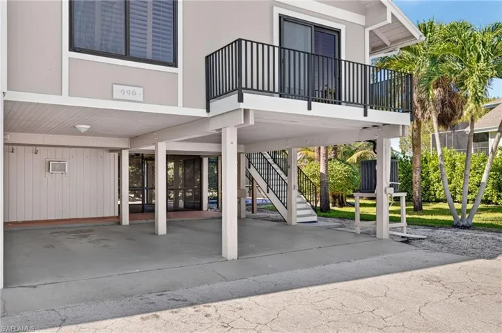 Covered ground-level parking area featuring a concrete floor, structural support columns, flush-mounted lighting, a staircase with metal railings, and a balcony with black metal balusters