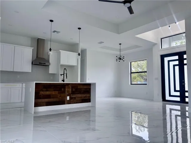 Kitchen featuring wall chimney exhaust hood, ceiling fan with notable chandelier, a raised ceiling, decorative light fixtures, and white cabinetry