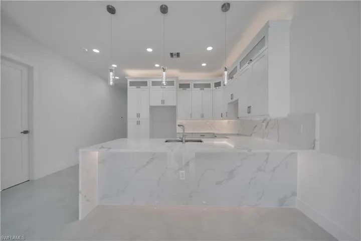 Kitchen featuring light stone countertops, a peninsula, white cabinetry, decorative light fixtures, and glass fronted cabinets
