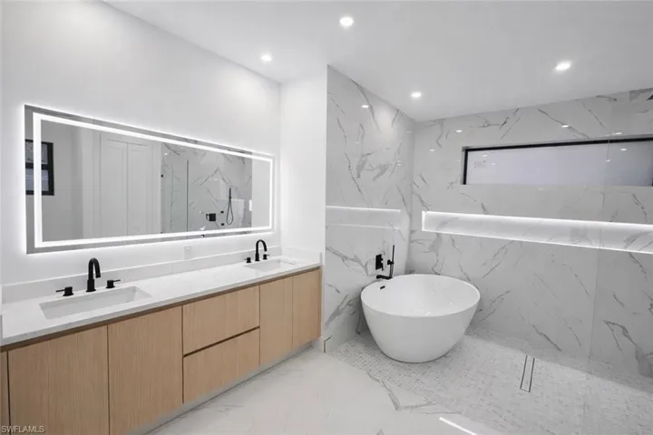 Full bath with stone wall, a freestanding bath, double vanity, recessed lighting, and light marble finish floors