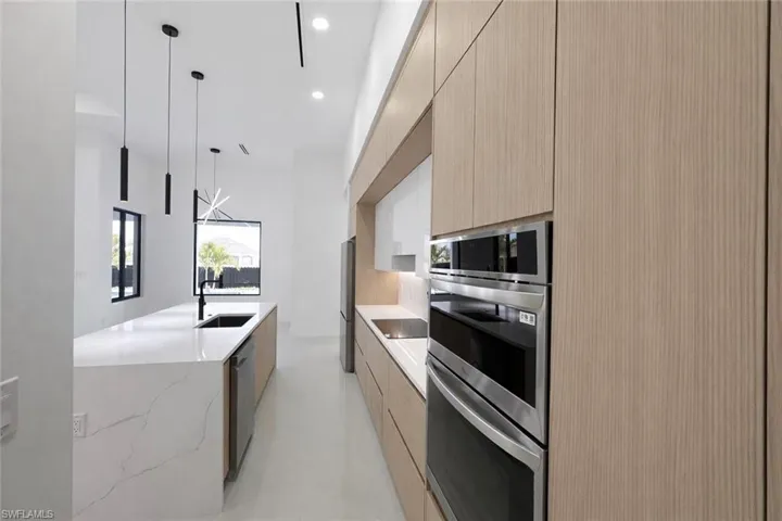 Kitchen with modern cabinets, stainless steel appliances, light wood finish cabinets, light stone counters, and hanging light fixtures