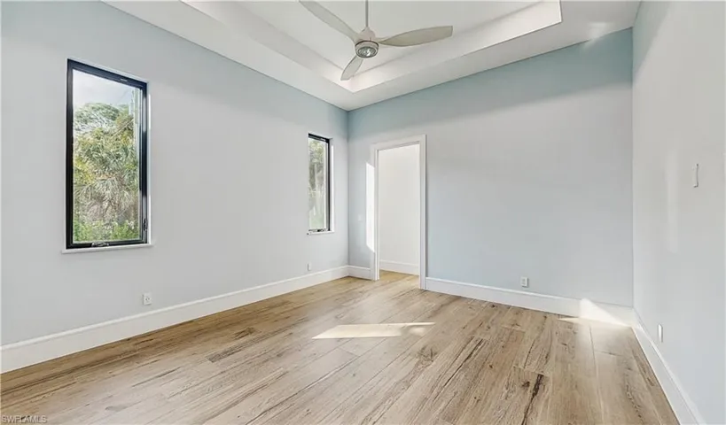 Empty room featuring a raised ceiling, ceiling fan, light wood-type flooring, and a wealth of natural light