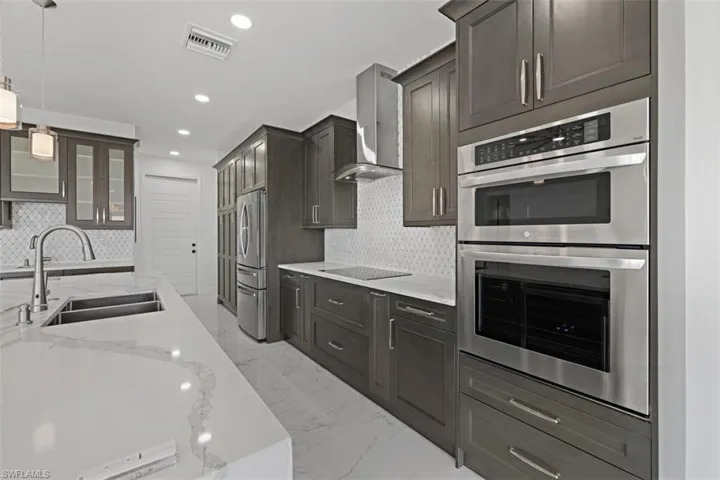 Kitchen featuring appliances with stainless steel finishes, glass insert cabinets, light stone counters, wall chimney exhaust hood, and light marble finish flooring