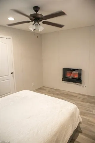 Bedroom with light wood-style floors, a glass covered fireplace, ceiling fan, and a decorative wall