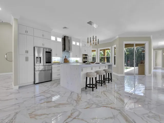 Kitchen with appliances with stainless steel finishes, wall chimney exhaust hood, light marble finish floors, a large island with sink, and white cabinets
