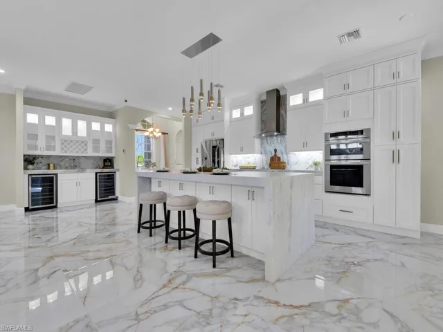 Kitchen featuring beverage cooler, wall chimney range hood, stainless steel double oven, light marble finish flooring, and crown molding