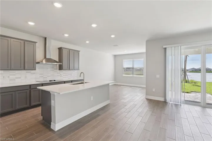 Kitchen with wood finished floors, gray cabinets, decorative backsplash, wall chimney range hood, and a kitchen island with sink