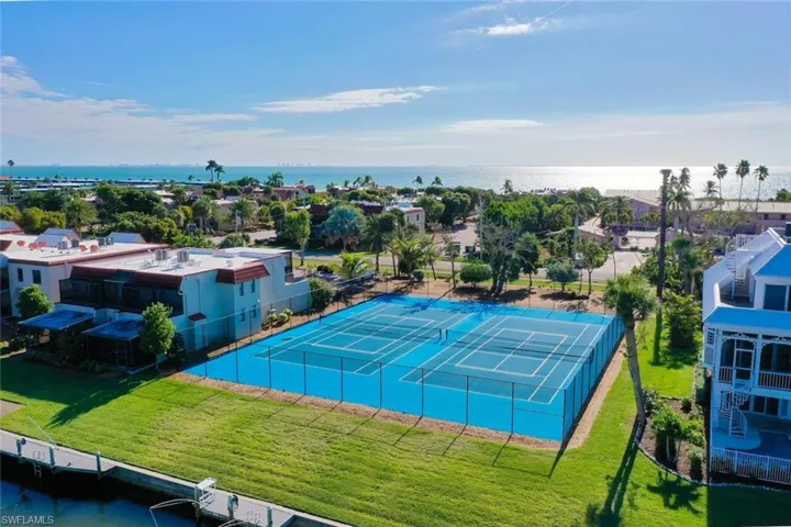 Blue tennis courts with surrounding fencing, adjacent to a waterway with boat docks, and visible water in the background