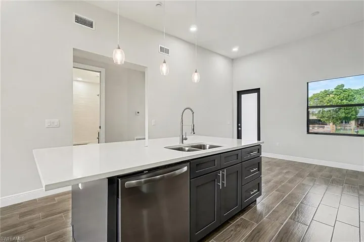 Kitchen with dishwasher, wood finish floors, hanging light fixtures, an island with sink, and light stone counters