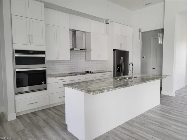 Kitchen featuring wall chimney range hood, an island with sink, stainless steel appliances, white cabinets, and light stone counters