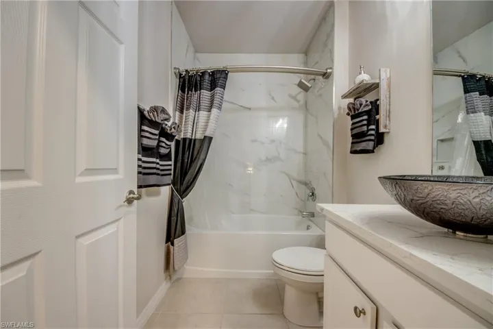 Bathroom featuring a built-in tub with a shower fixture, wall-mounted marble-finish tile surround, and a vanity with a vessel sink