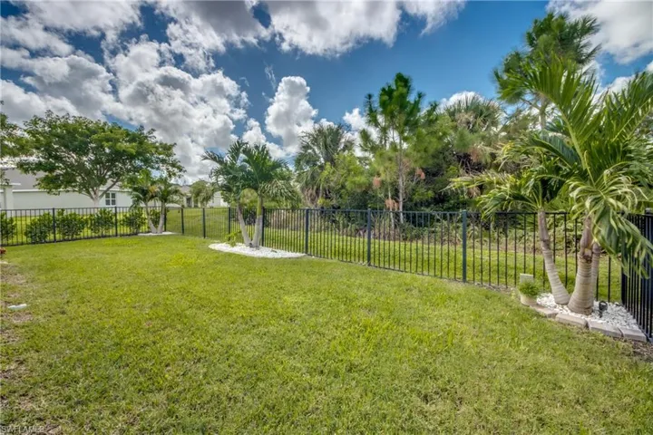 Expansive grassy backyard featuring a black metal fence, mature trees, and palm trees with white rock landscaping