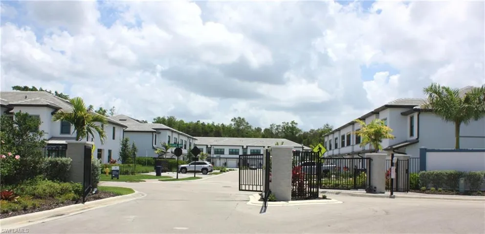 Gated community entrance featuring modern stucco homes with tile roofs, lush landscaping, and mature palm trees