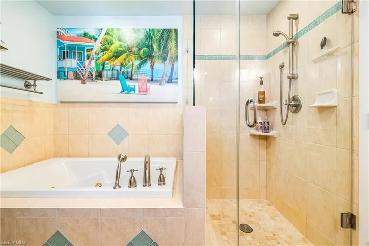 Jetted soaking tub with deck-mounted handheld sprayer, frameless glass-enclosed shower, wall-mounted adjustable showerhead, and neutral tile wall surrounds with decorative accent inserts