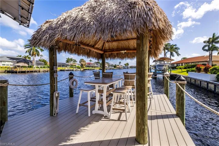 Waterfront boat dock featuring a synthetic deck surface, wood-post pilings, rope railings, and a thatch-roof palapa