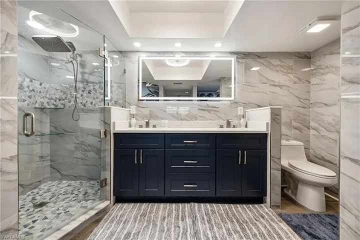 Contemporary bathroom featuring a dark blue dual vanity with a white countertop, an illuminated mirror, a glass-enclosed shower with a rain showerhead, and light-toned wall tiling