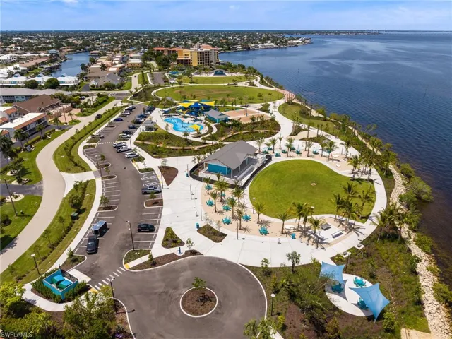 Expansive waterfront park area featuring a large green space, paved pathways, a water play area, and an open-air pavilion