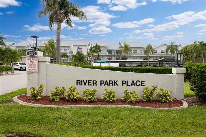 Community entryway featuring a stucco monument sign with decorative landscaping, established palm trees, and exterior lighting fixtures