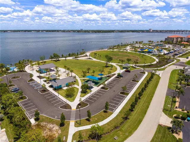 Expansive waterfront park area featuring manicured green spaces, paved walking paths, covered picnic shelters, a children's play area, and dedicated parking facilities