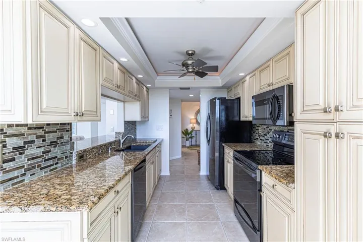 Kitchen with light stone counters, black appliances, a tray ceiling, ceiling fan, and decorative backsplash