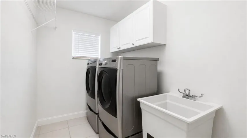 Dedicated laundry area featuring a utility sink with dual-handle faucet, overhead white cabinetry, window with horizontal blinds, and a wire storage shelf