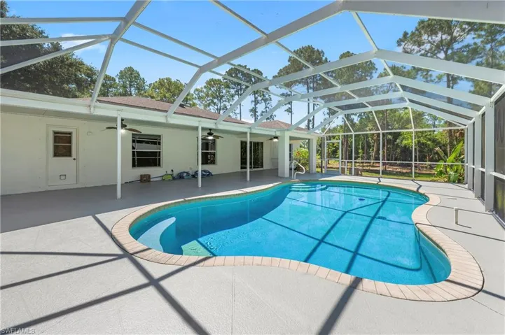 Curved swimming pool within a screened enclosure featuring a spacious concrete deck and an exterior wall with multiple access points and ceiling fans