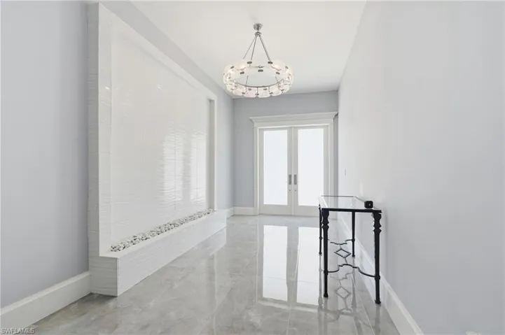 Hallway featuring a built-in water wall with textured white tile and decorative stones, double-door entry, overhead chandelier, and polished stone tile flooring