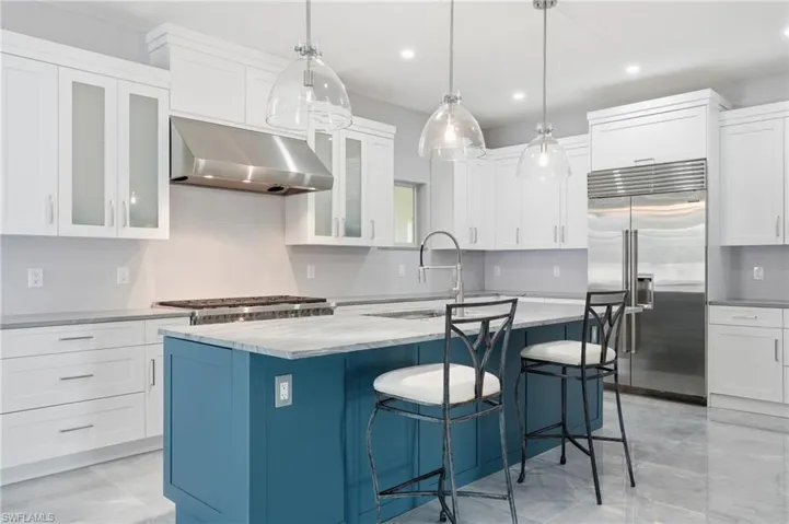 Kitchen featuring a blue island with a contrasting stone countertop, clear pendant lighting, stainless steel appliances, white cabinetry with some frosted glass doors, and light-toned tile flooring