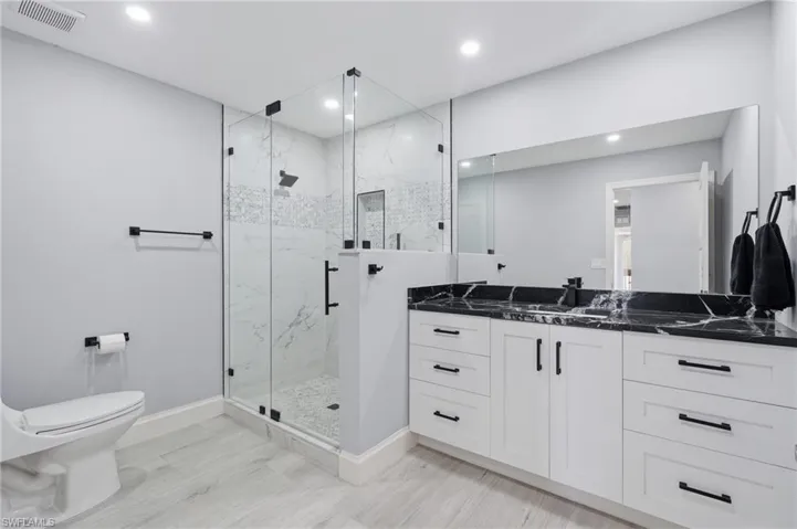 Contemporary bathroom featuring a spacious glass-enclosed shower with white marble-look tile, a dual vanity with a black stone countertop, white shaker-style cabinetry, and light wood-finish flooring