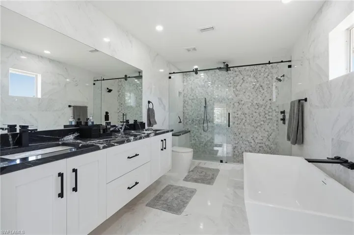 Spacious bathroom featuring a dual vanity with black countertops, a freestanding soaking tub, a walk-in shower with mosaic tile, and marble-look tile flooring and walls