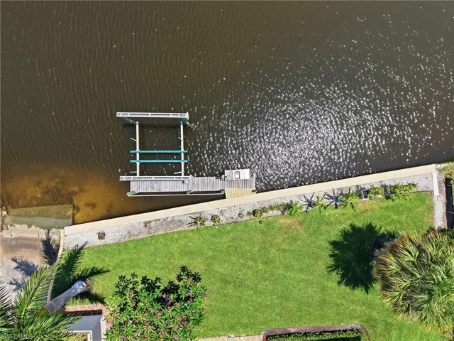 Waterfront property featuring a private boat lift, a boat dock, a concrete seawall, a manicured lawn, and mature tropical landscaping
