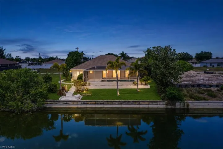 Waterfront property featuring a screened-in pool area, mature palm trees, manicured lawn, and a concrete waterside retaining wall