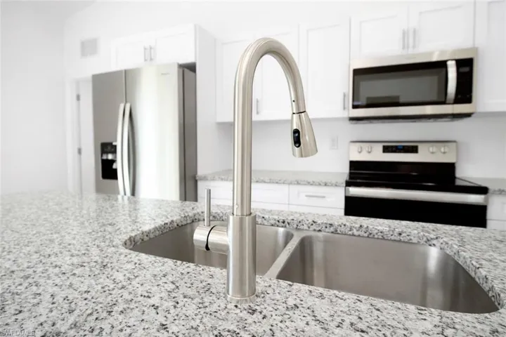Kitchen featuring a double stainless steel sink with a pull-down faucet, granite countertops, white cabinetry, and stainless steel appliances including a refrigerator, range, and microwave