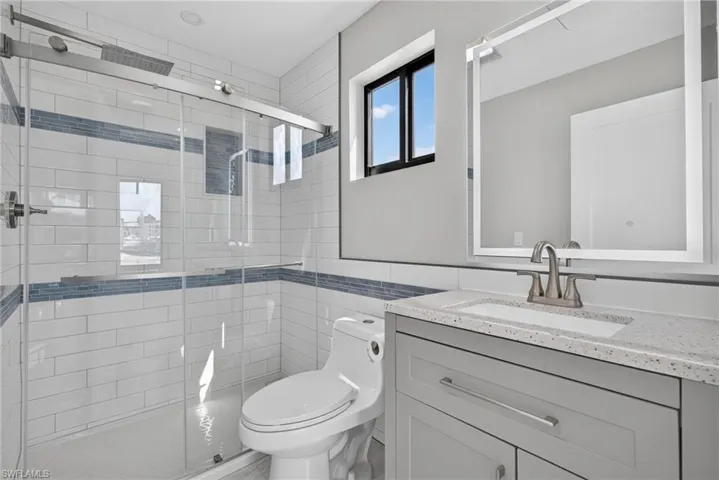 71 Mango Unit 1 Contemporary bathroom featuring a glass-enclosed shower with subway tile surround and integrated niche, a single-sink vanity with light gray cabinetry and a speckled countertop, a rectangular mirror, and a black-framed window