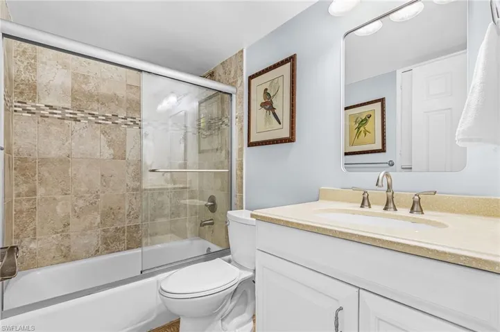 Tub with sliding glass doors, stone-look tile surround with decorative accent band, white vanity with stone-finish countertop, and wall-mounted mirror