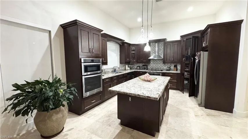 Kitchen with dark brown cabinets, wall chimney exhaust hood, stainless steel appliances, a kitchen island, and hanging light fixtures