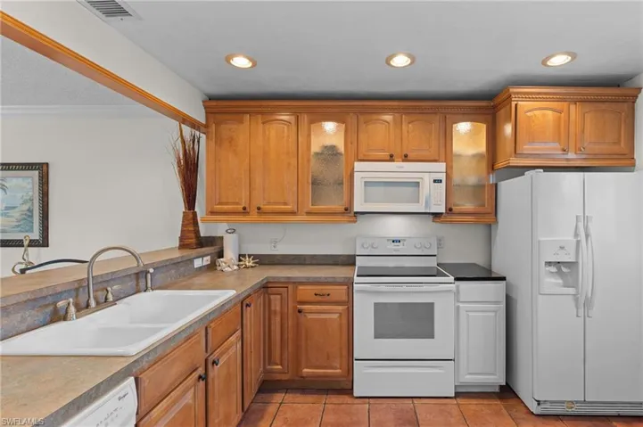Kitchen with white appliances, glass insert cabinets, brown cabinets, dark countertops, and recessed lighting