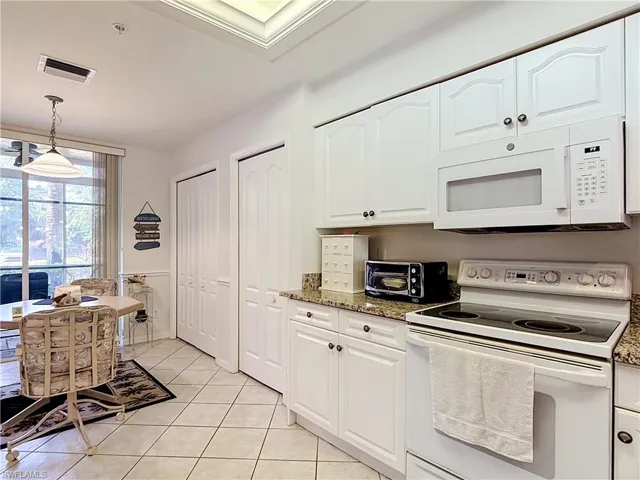 White cabinetry with dark hardware, stone-finish countertops, stainless steel appliances, tiled flooring, and a large window with vertical blinds