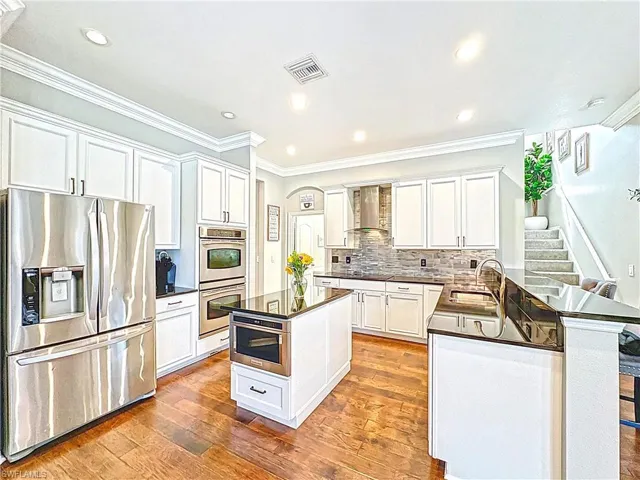 Kitchen with stainless steel appliances, dark countertops, wall chimney range hood, hardwood / wood-style flooring, and ornamental molding