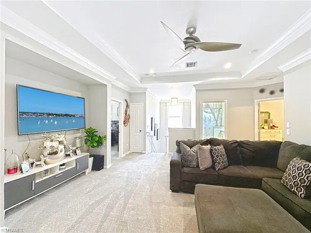 Living area with a raised ceiling, a ceiling fan, light carpet, and ornamental molding