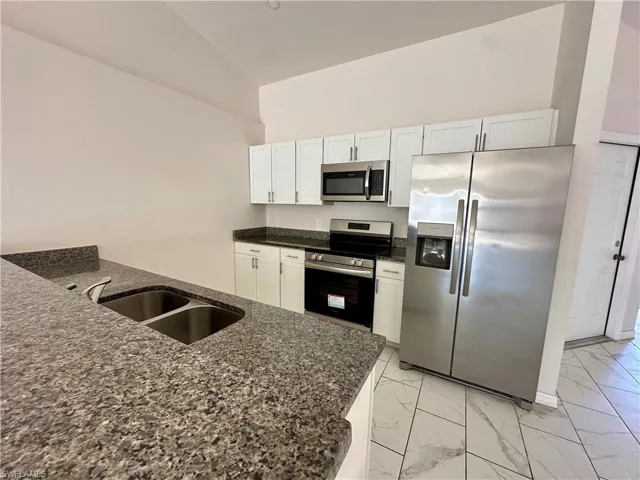 Kitchen with stainless steel appliances, white cabinets, light marble finish flooring, dark stone countertops, and vaulted ceiling
